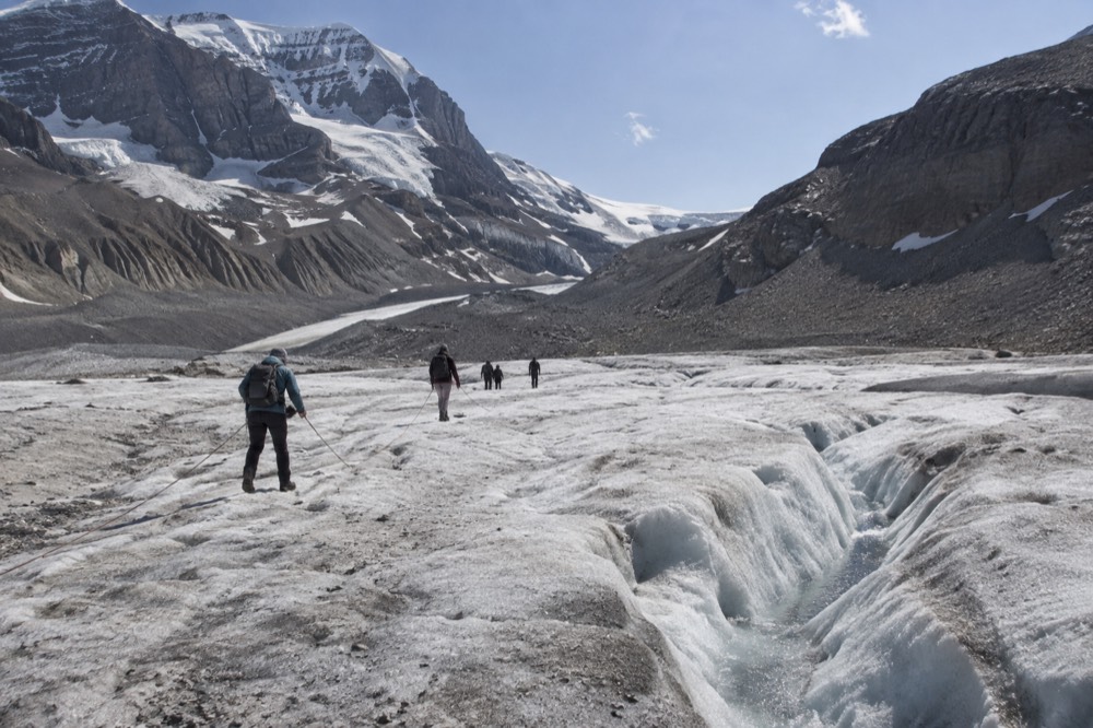 A group of hikers in crampons walking across a crevassed glacier tongue under a bright sky