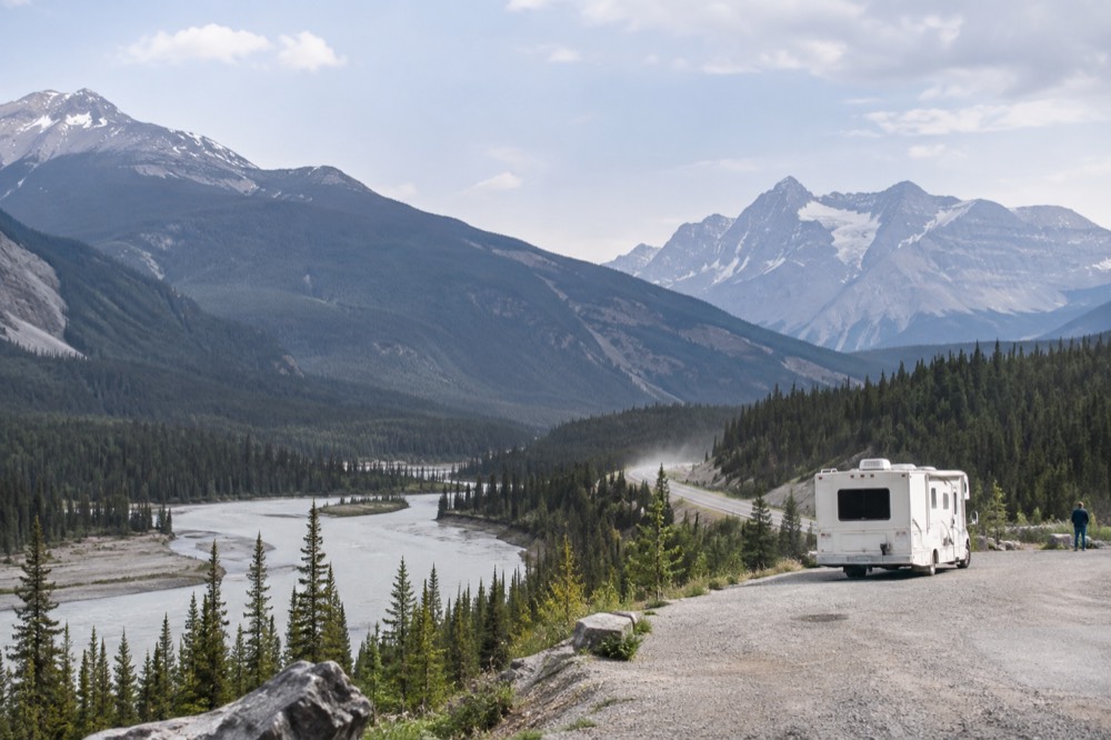 A winding mountain road with a glacier-fed river running alongside it under tall peaks
