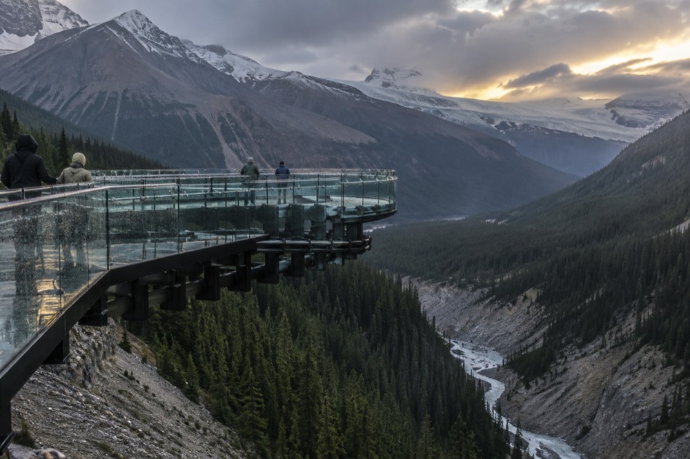 A curved glass-floored walkway extending over a deep mountain valley with a river far below