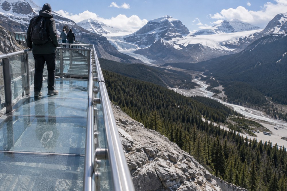 A glass-floored walkway suspended along a cliff edge above a deep forested river valley