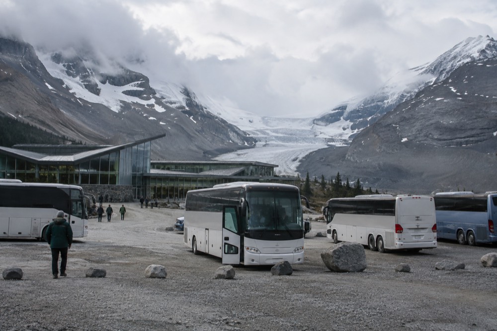 The Columbia Icefield Discovery Centre building at the foot of the Athabasca Glacier, with tour buses in the parking area and the icefield rising behind.