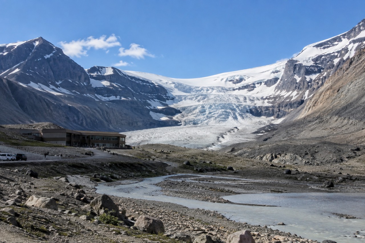 The Athabasca Glacier flowing down from the Columbia Icefield under a blue sky, with the Icefield Centre building in the valley below.