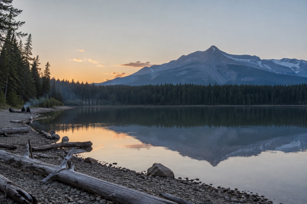 A calm lake reflecting mountains and pine forest under a clear sky at dusk