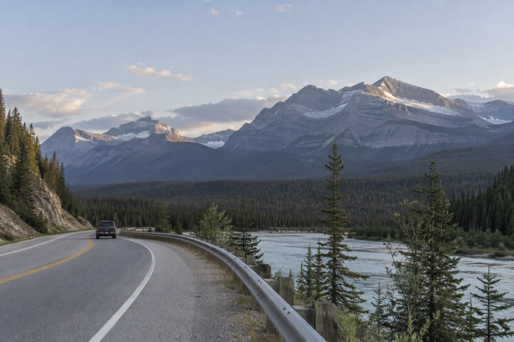 A winding road through a mountain valley beside a river with rocky peaks behind