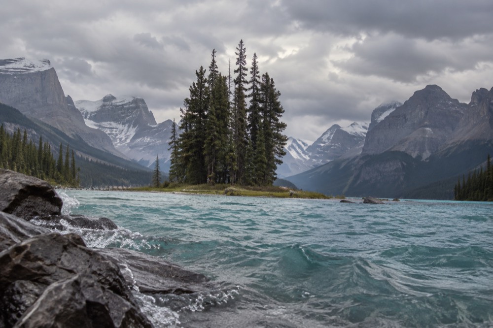 Turquoise lake water between forested slopes with distant snow-capped mountains under scattered clouds