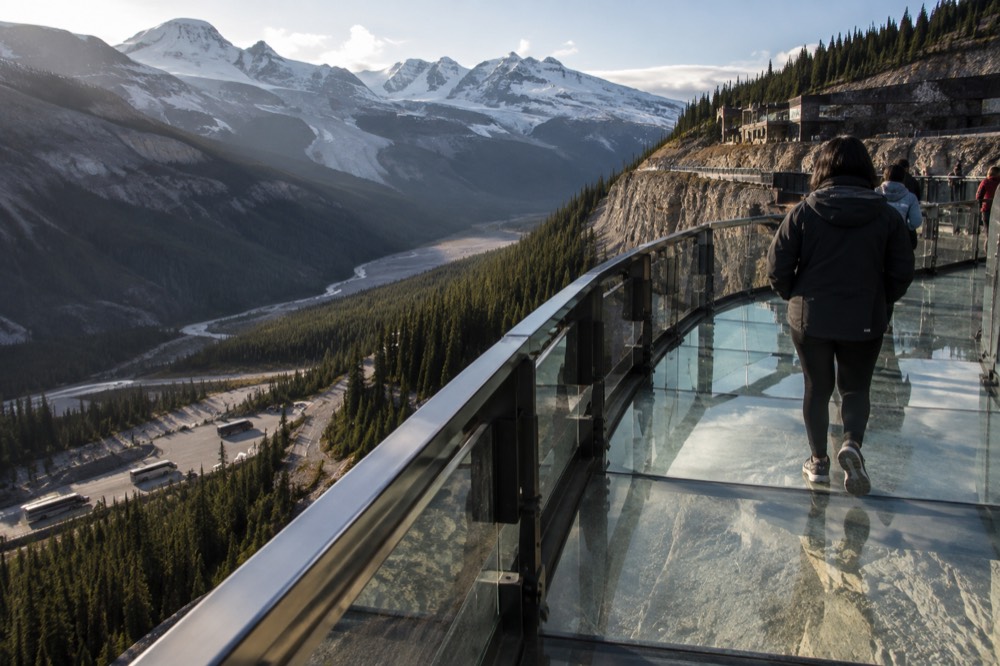 Cliff-edge walkway with a glass floor section overlooking a wide glacial valley and distant mountain peaks