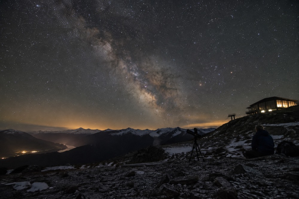 Night sky filled with stars above a dark mountain silhouette and a small lit building