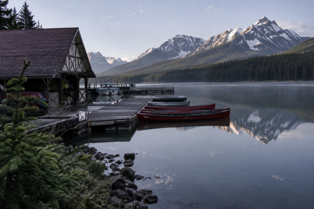 A calm lake reflecting mountains in early morning light with a wooden boathouse dock