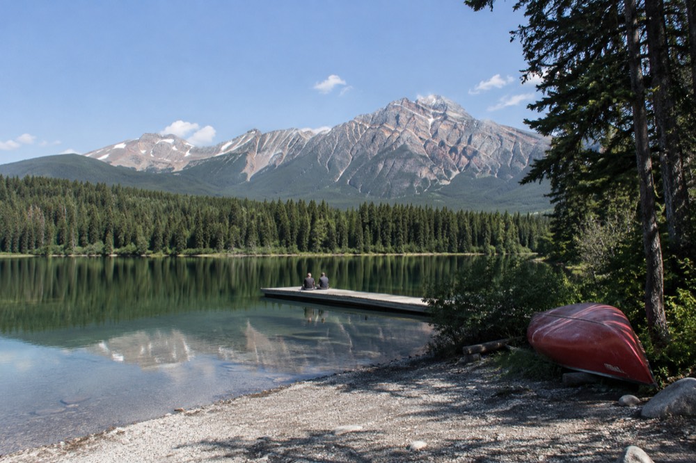 A calm glacial lake reflecting pine-covered mountains under a clear summer sky