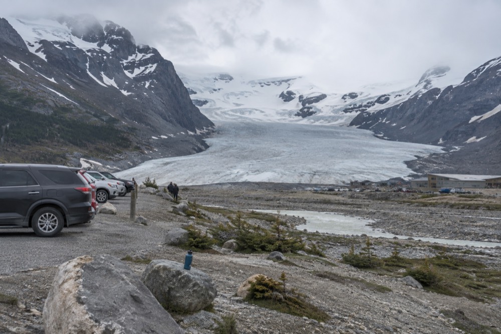 A glacier tongue flowing down between dark rock walls with a parking area and small buildings near its edge