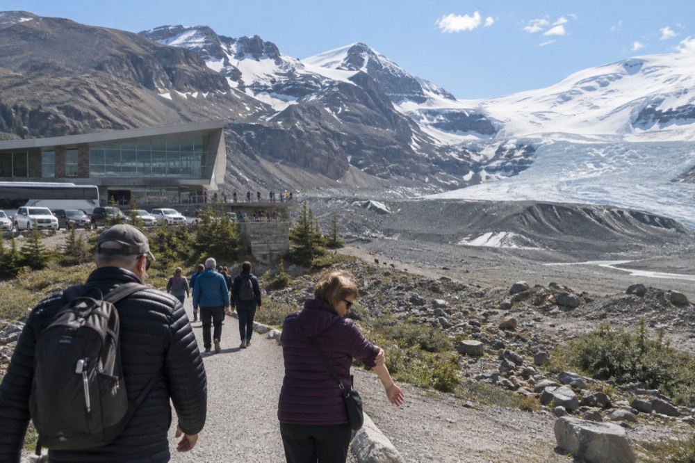 Visitors in jackets and sunglasses walking a gravel path beside a glacier forefield