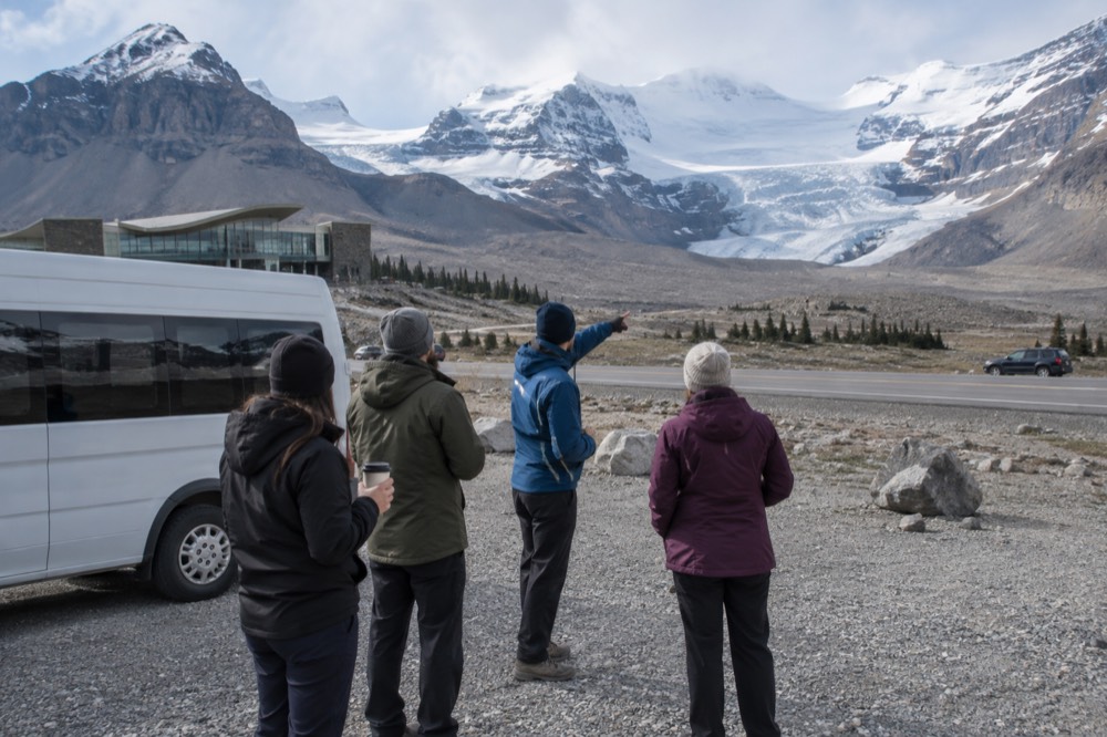 A small tour van parked at a glacier viewpoint with a guide speaking to passengers
