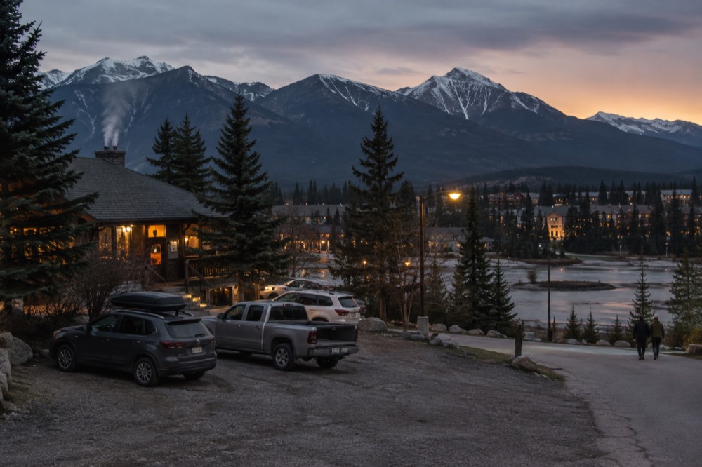 A mountain town beside a river at dusk with warm lights and peaks behind