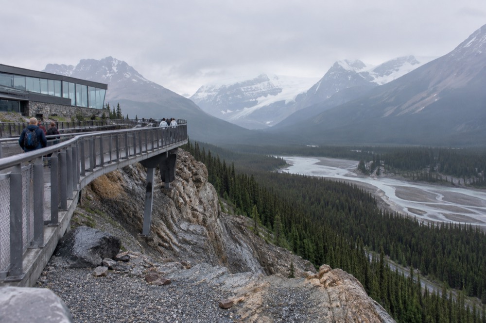 A curved walkway clinging to a tall cliff face above a wide forested valley with a braided river below