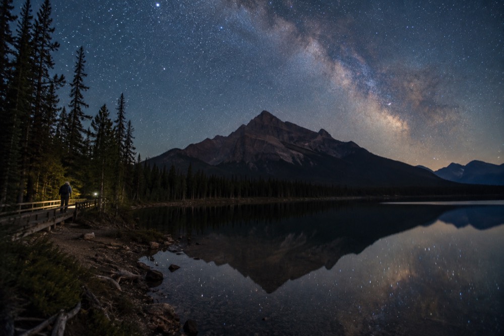 The Milky Way arching over a dark mountain silhouette with a lake in the foreground