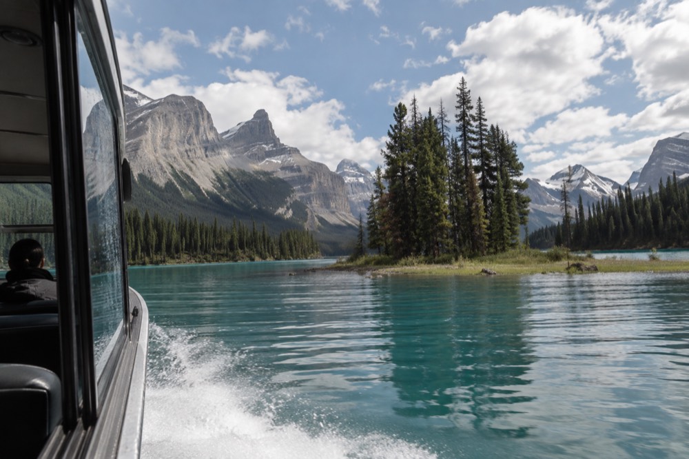 A boat crossing a long blue glacial lake toward a small forested island with mountains behind