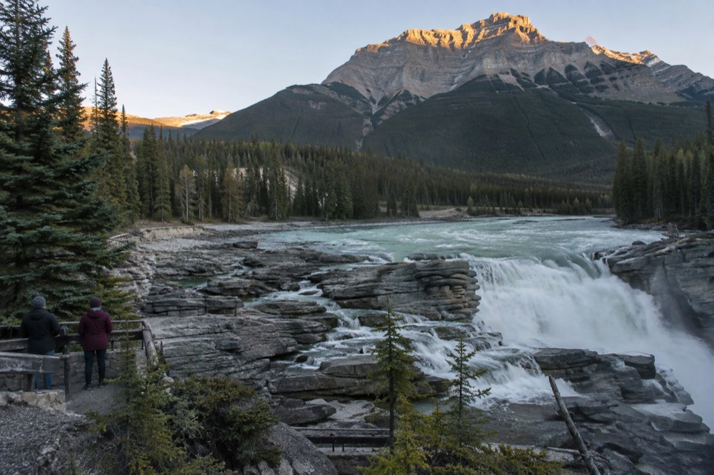 A quiet asphalt highway curving beside a turquoise river with tall snow-capped mountains rising on both sides