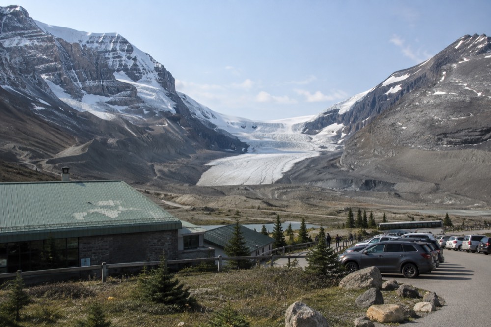 A broad glacier tongue flowing down between dark rock walls with a parking area and small buildings at its base