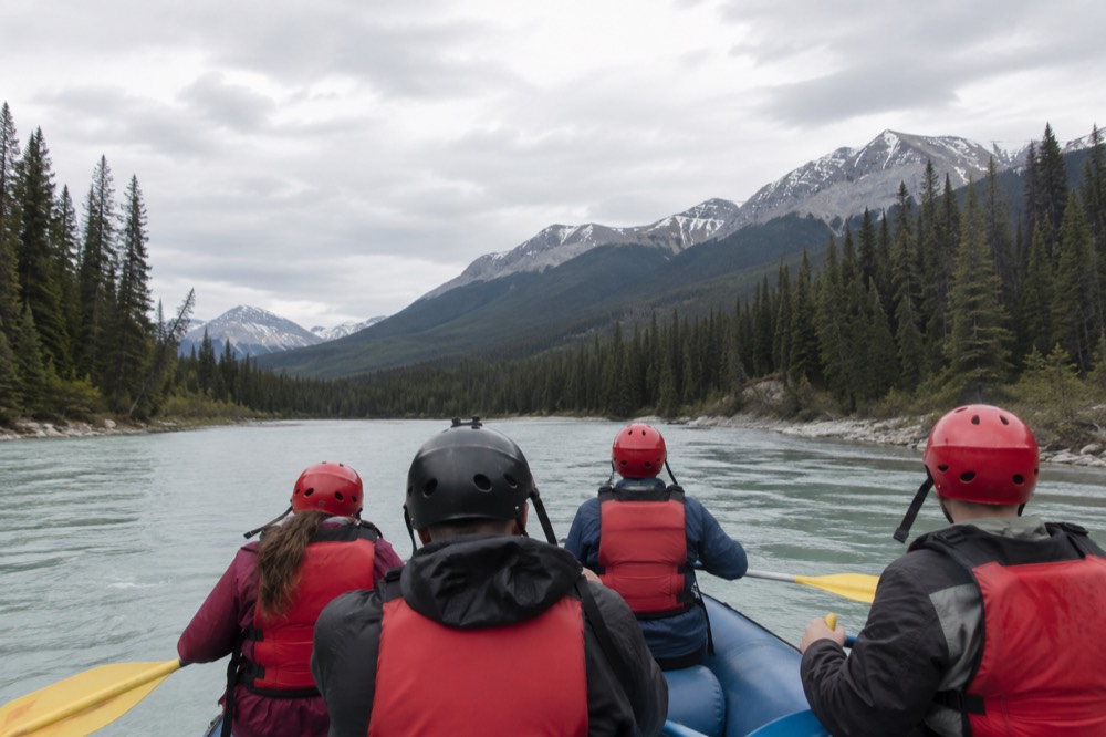 A raft with paddlers in life jackets floating down a wide turquoise river between forested mountains