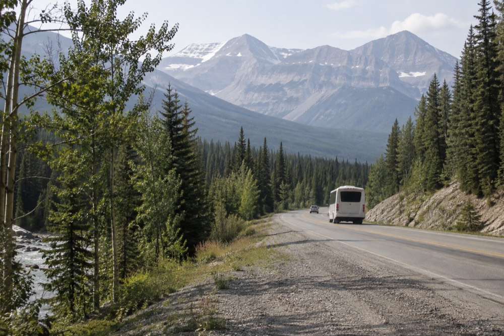 A small tour bus on a forest highway heading toward distant peaks