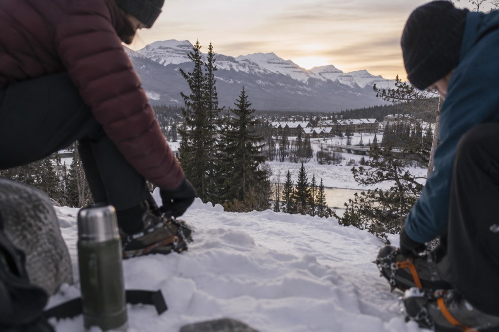 Winter hikers in insulated jackets and boots adjusting boot cleats on a snowy trail