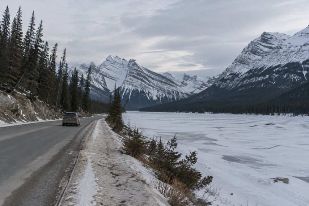 A winding mountain road beside a frozen river with snow-covered peaks beyond