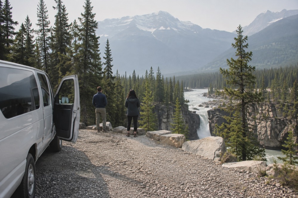 Tour van parked at a roadside viewpoint above a turquoise river and forested valley