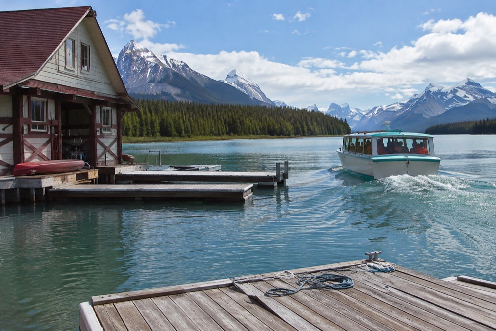 A wooden tour boat carrying passengers across the deep green water of Maligne Lake with mountains rising on both sides
