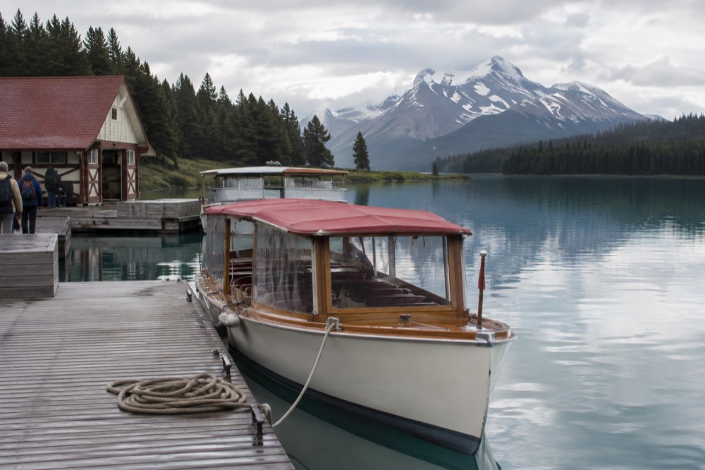 A small wooden boat with a red canopy moored at a dock on a glassy lake surrounded by forested shores and mountain peaks