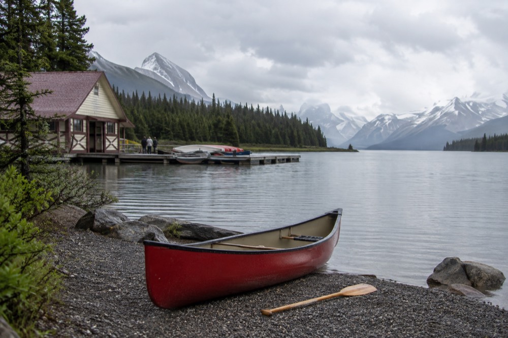 A red canoe pulled up on a rocky shore with a mountain lake stretching into the distance under a cloudy sky