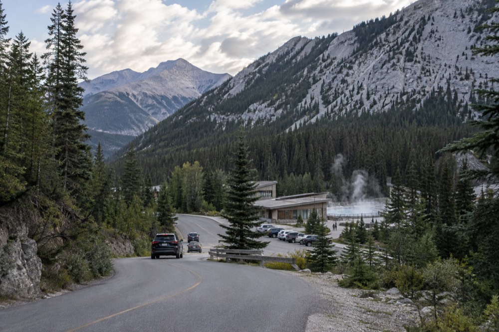 Winding mountain road through forested valley with high snow peaks ahead under scattered clouds