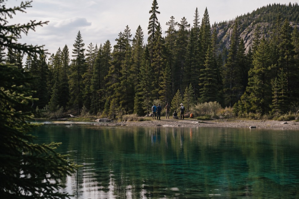 A small group of hikers listening to a guide beside a still lake with forested hills beyond
